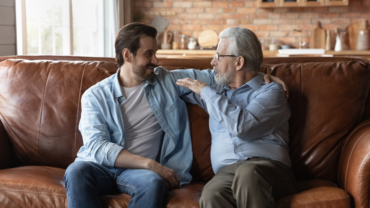 Adult son having a warm conversation with his elderly father on a couch, illustrating how to talk to your elderly parents about assisted living.