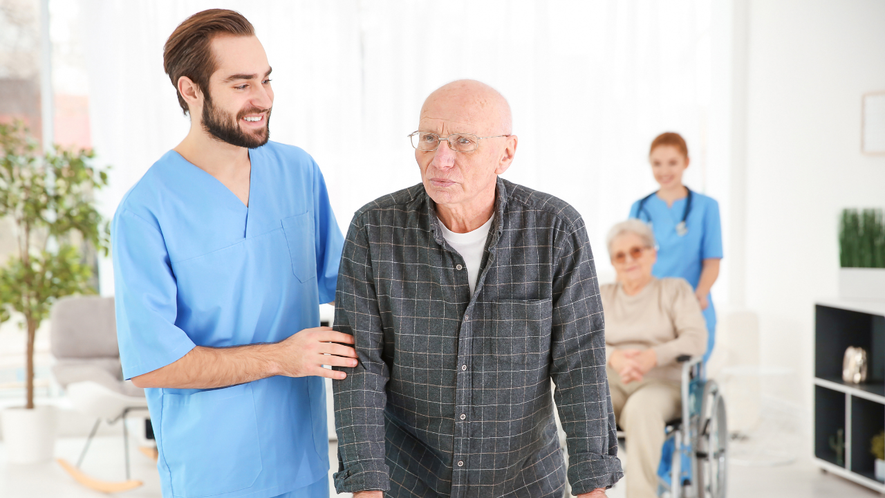 Caregiver assisting an elderly man with walking in a senior care facility, illustrating the difference between assisted living and skilled nursing.