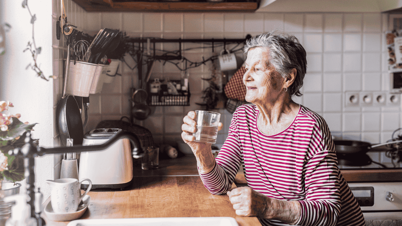 Senior woman in her kitchen drinking water, representing questions like does the state pay for elderly care support.